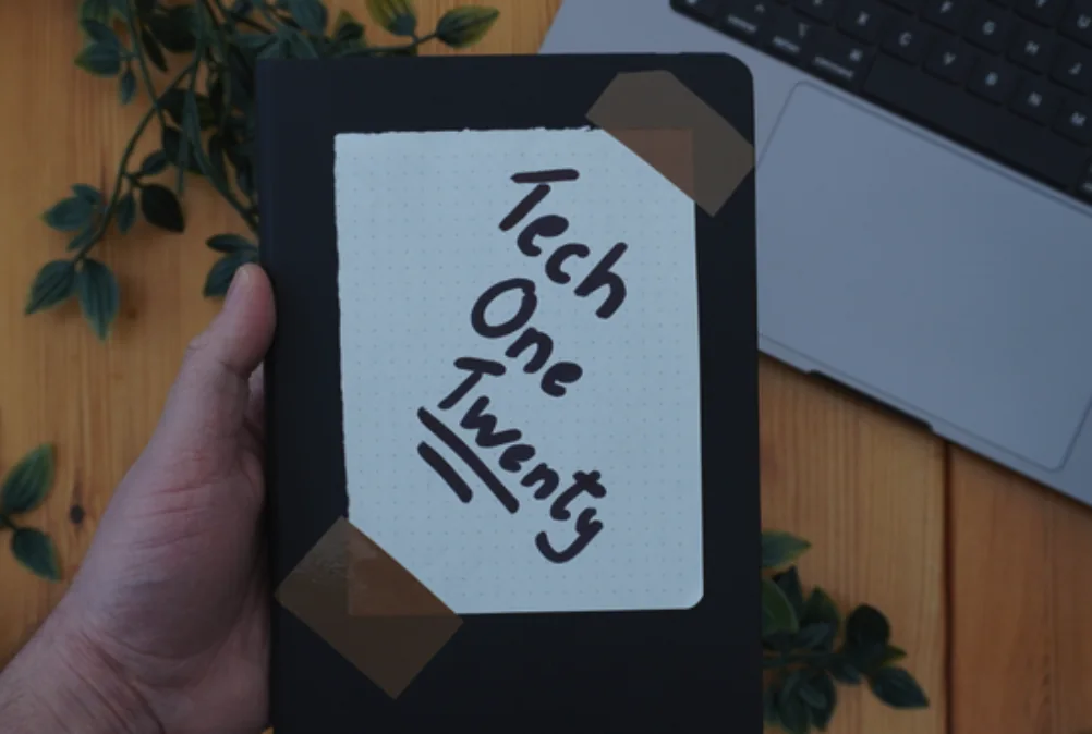 A hand holding a copy of Tech One Twenty on a desk beside a laptop